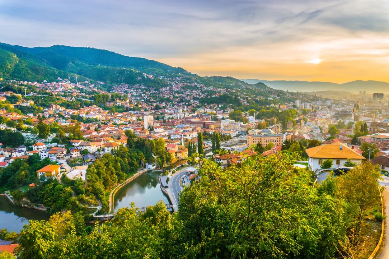 Aerial view of Sarajevo city nestled in green mountains at sunset with a river bend.