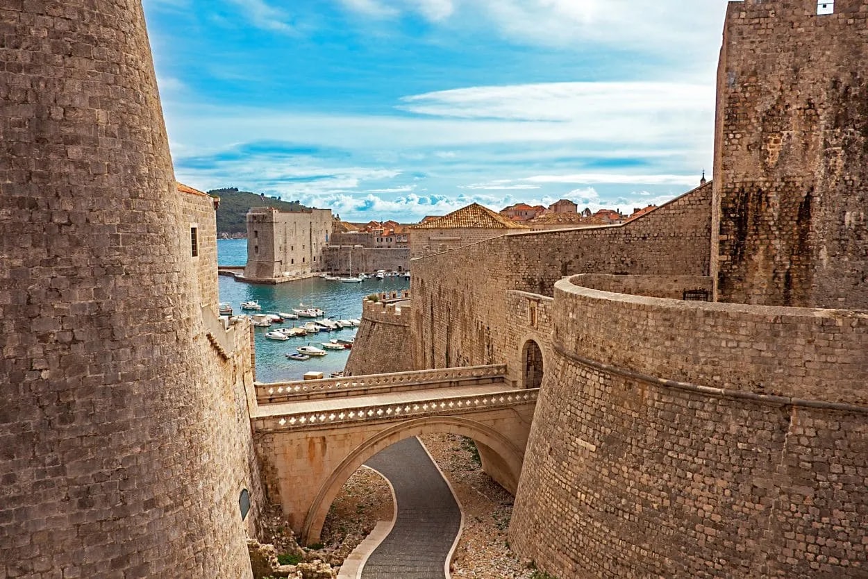 Fort walls of Dubrovnik with stone bridge overlooking a small harbor with boats and blue sky.