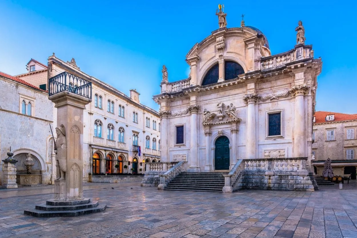 Baroque church and stone column in the Old Square in Dubrovnik under a clear blue sky.