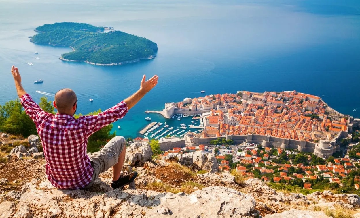 Man with arms raised overlooking Dubrovnik old town, harbor, and Lokrum Island from Mount Srd.