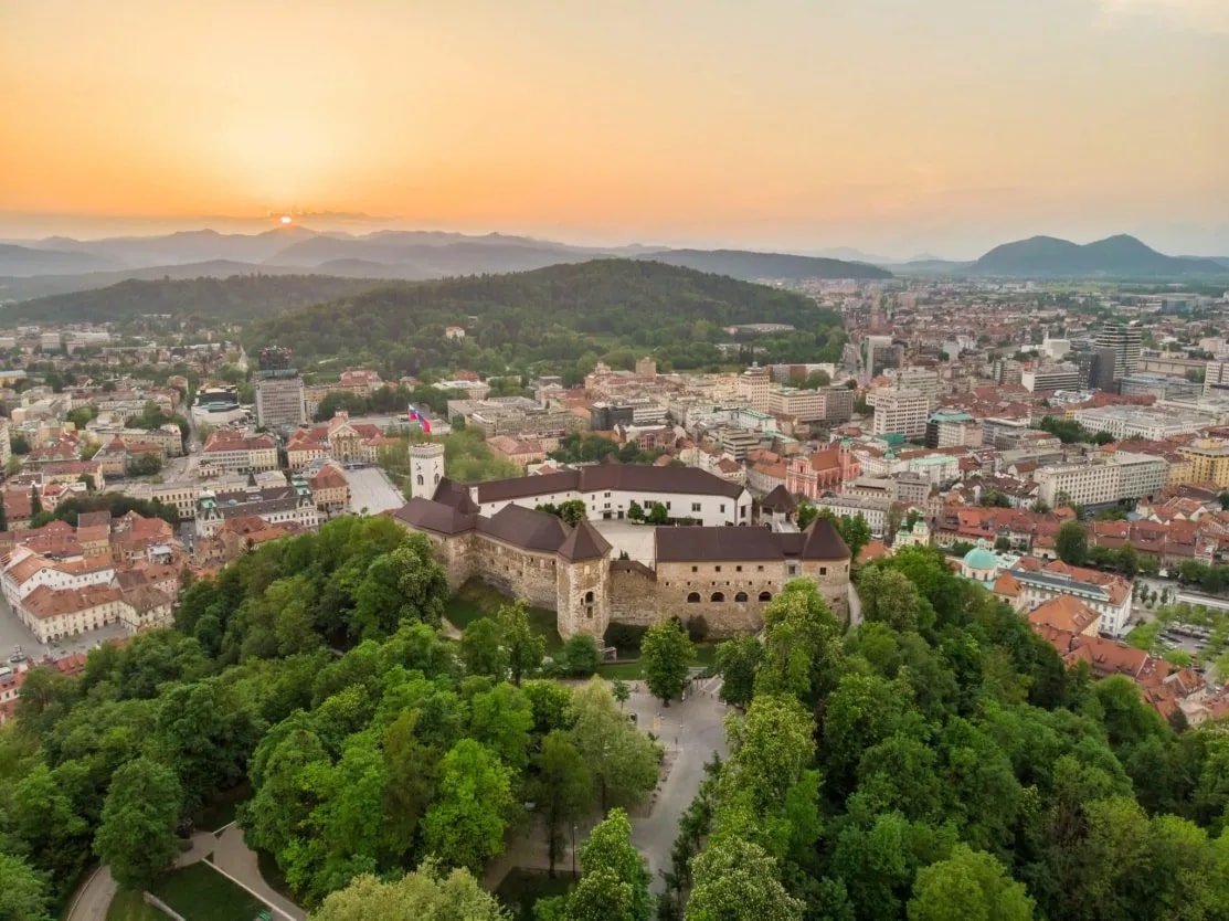 Ljubljana Castle on a wooded hill overlooking the city at sunset with distant mountains.