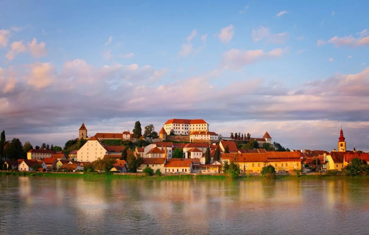 Ptuj historic town with castle on hill reflected in river under colorful sunset sky.