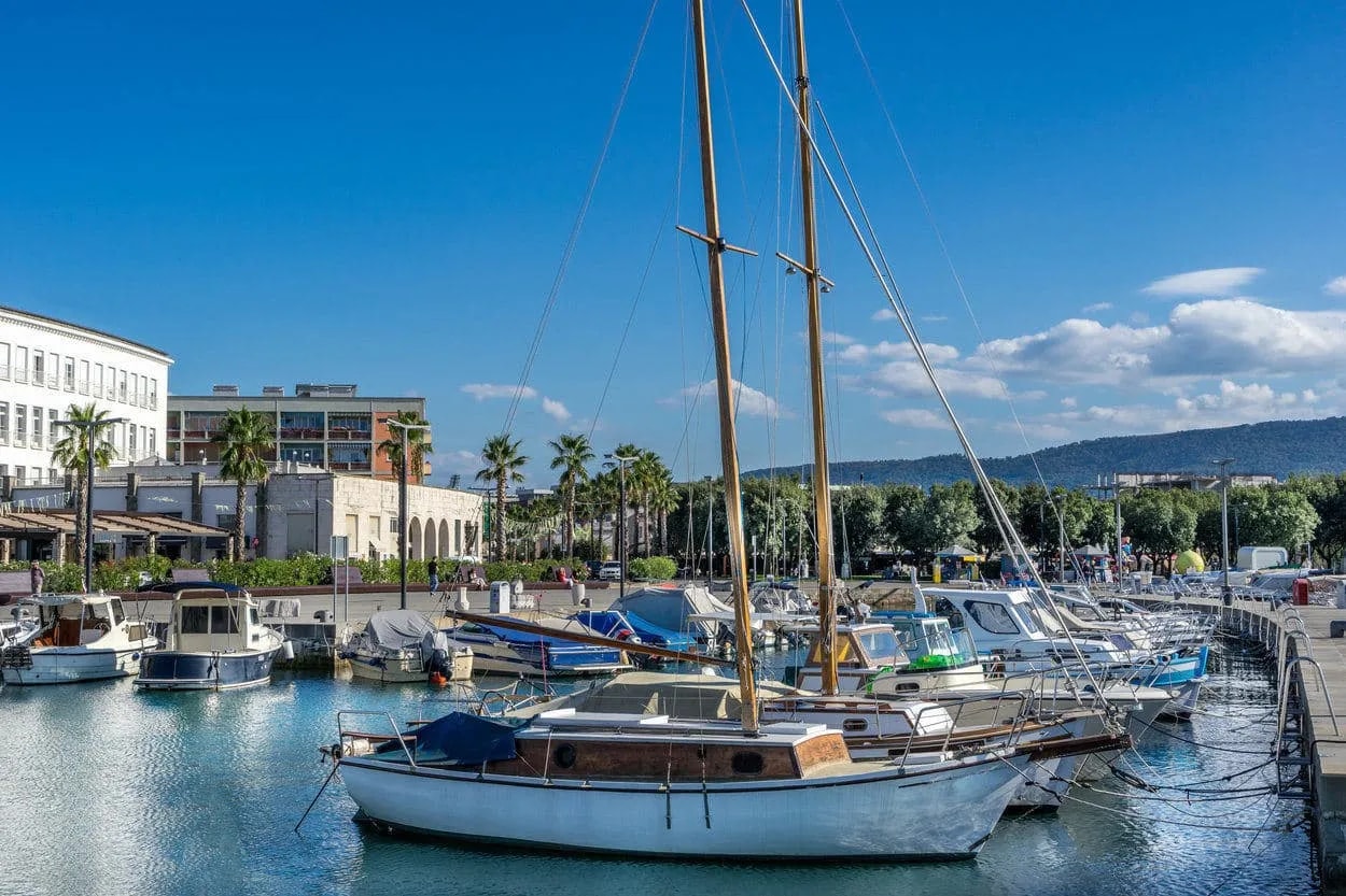 Sailboats and motorboats docked in the harbor of Koper under a bright blue sky with distant hills.