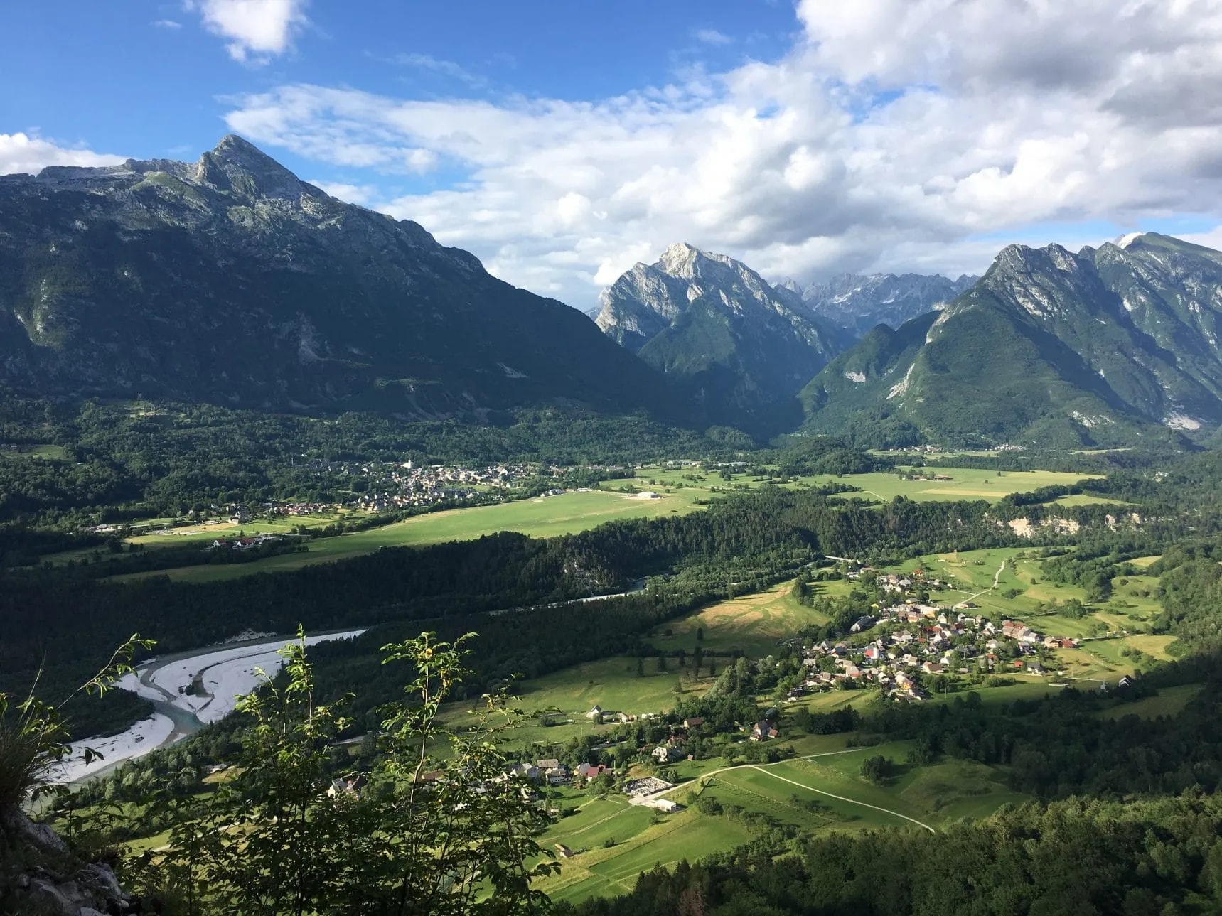 Panorama of Bovec valley with green fields, winding river, and Julian Alps mountains under a cloudy sky.