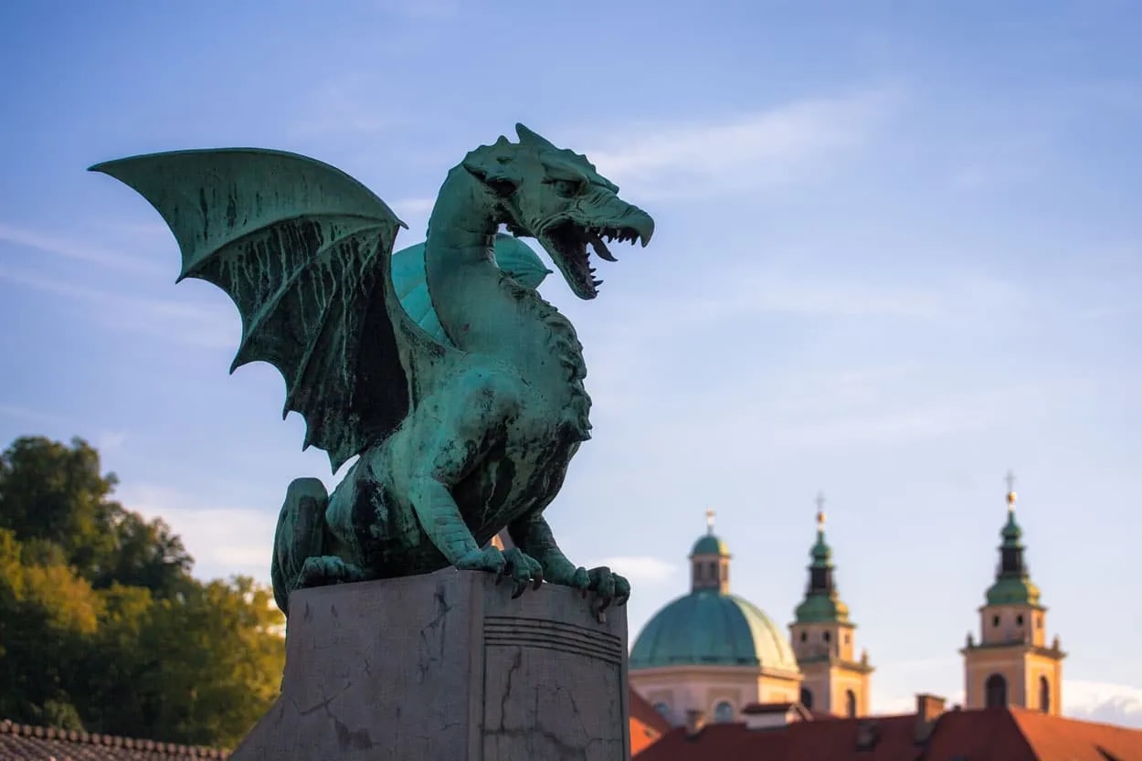 Verdigris dragon statue on pedestal with Ljubljana buildings in background.