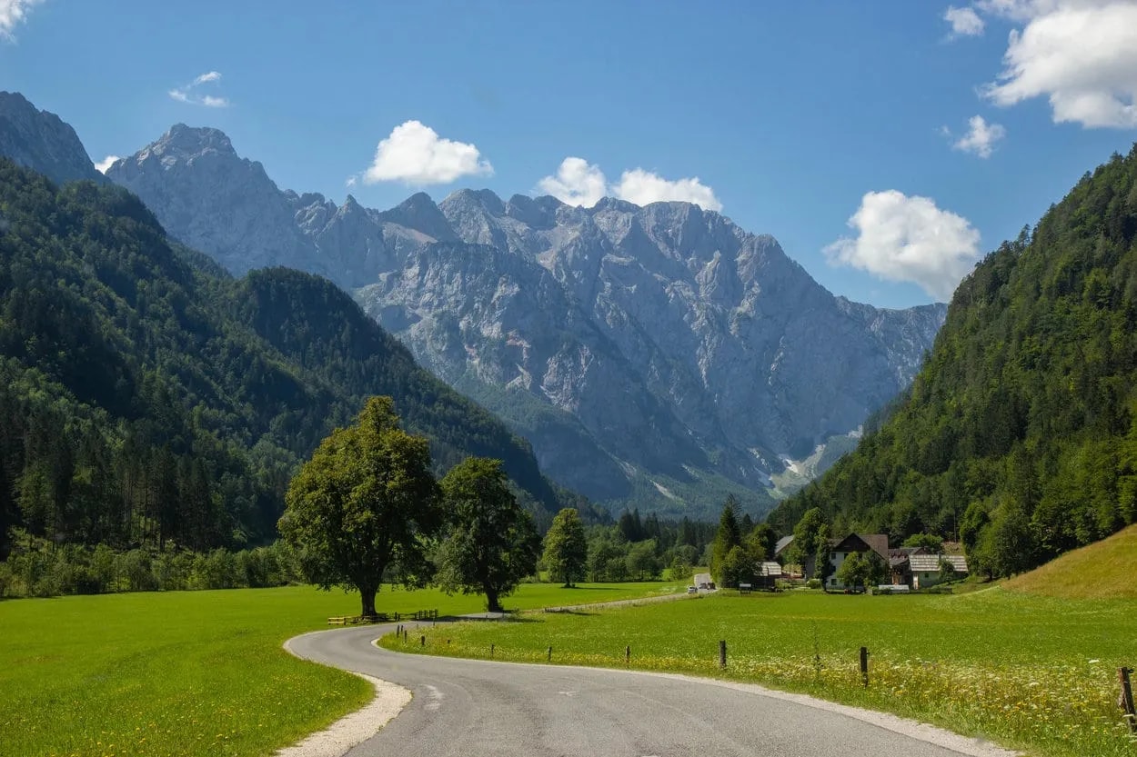 Winding road through green valley toward Logar Valley mountains under blue sky.