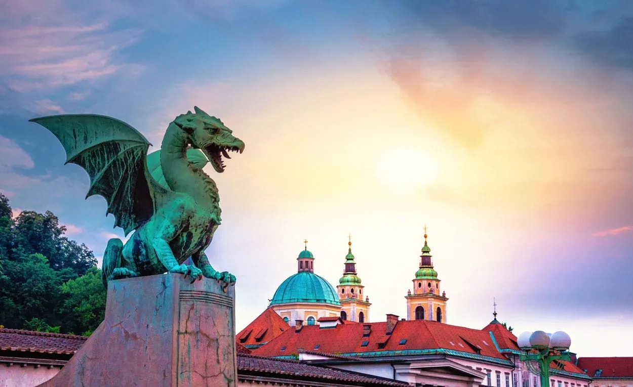 Green dragon statue on Dragon Bridge with Ljubljana cityscape and sunset sky.