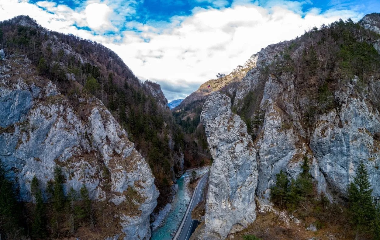 Road through a steep mountain gorge with a turquoise river and tall rock formations.