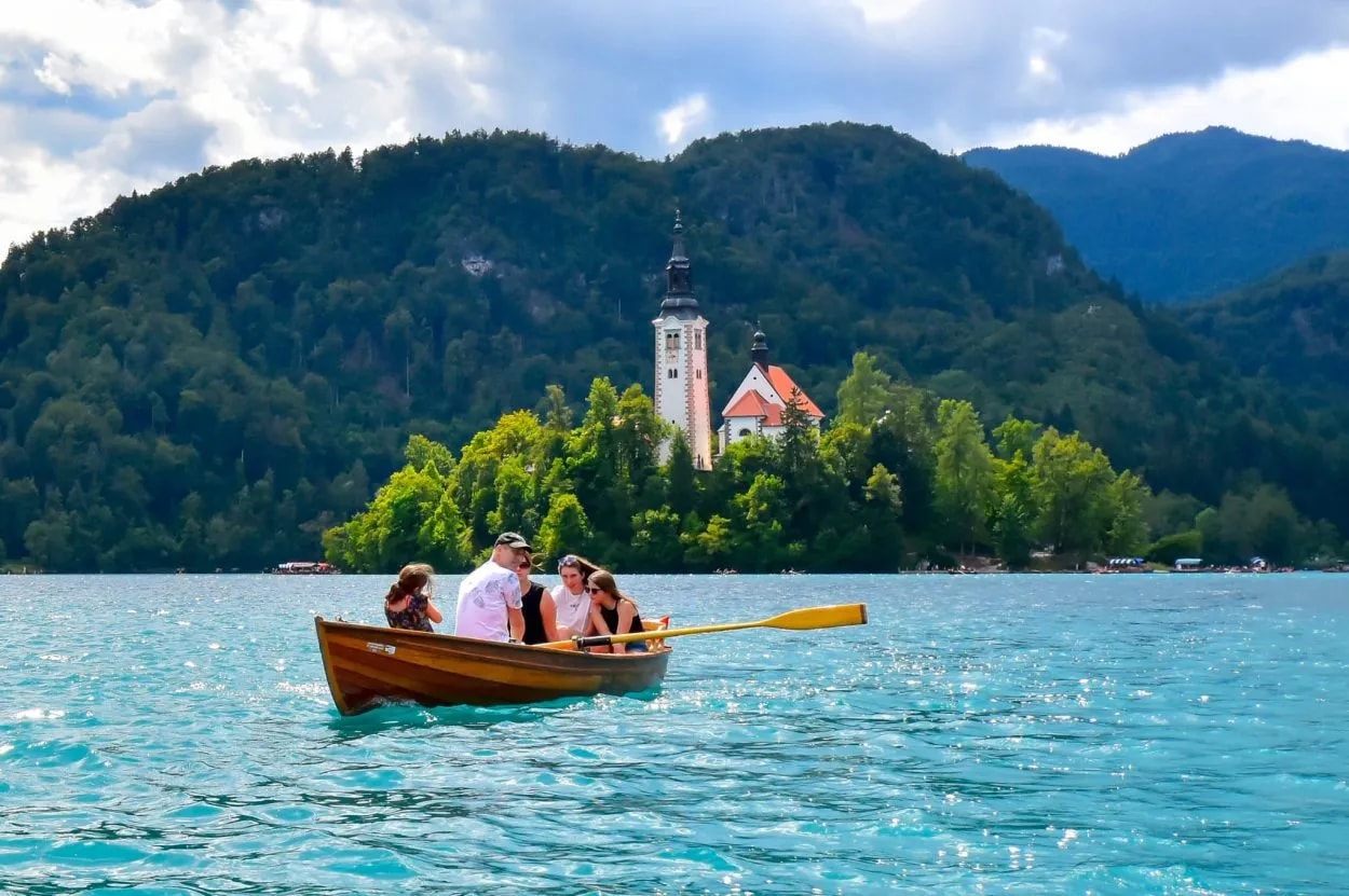 Rowboat with people on Lake Bled approaching the island church with forested mountains.