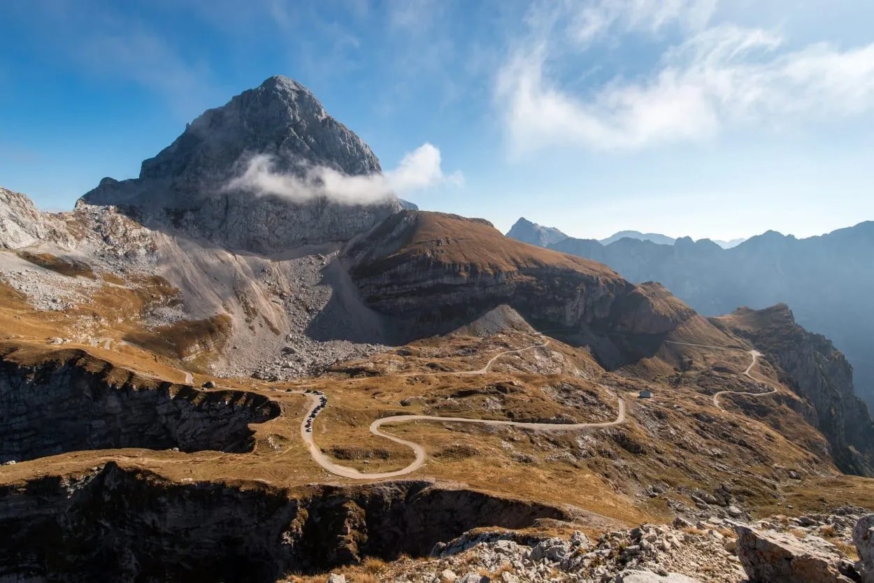 Winding mountain road ascending past rocky slopes toward a large peak under a blue sky.