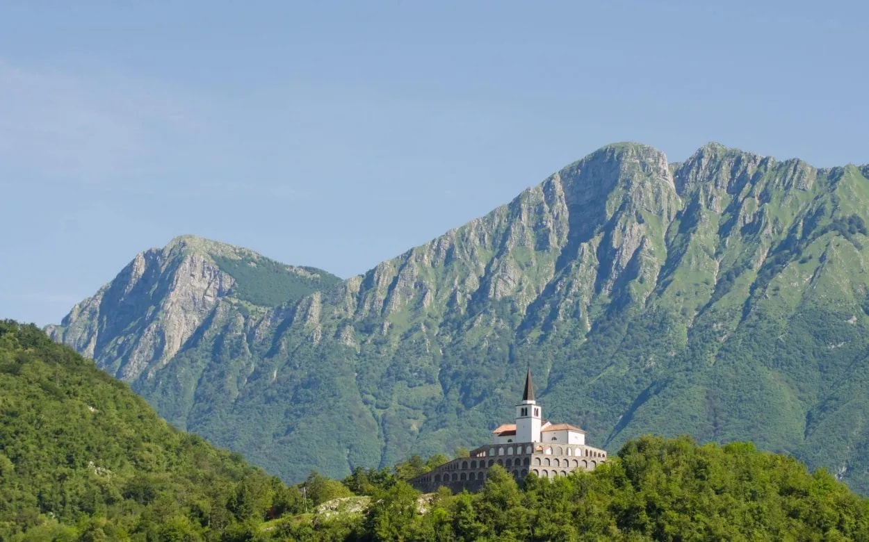 Church with a steeple on a green hill against rugged, forested mountains in Kobarid.