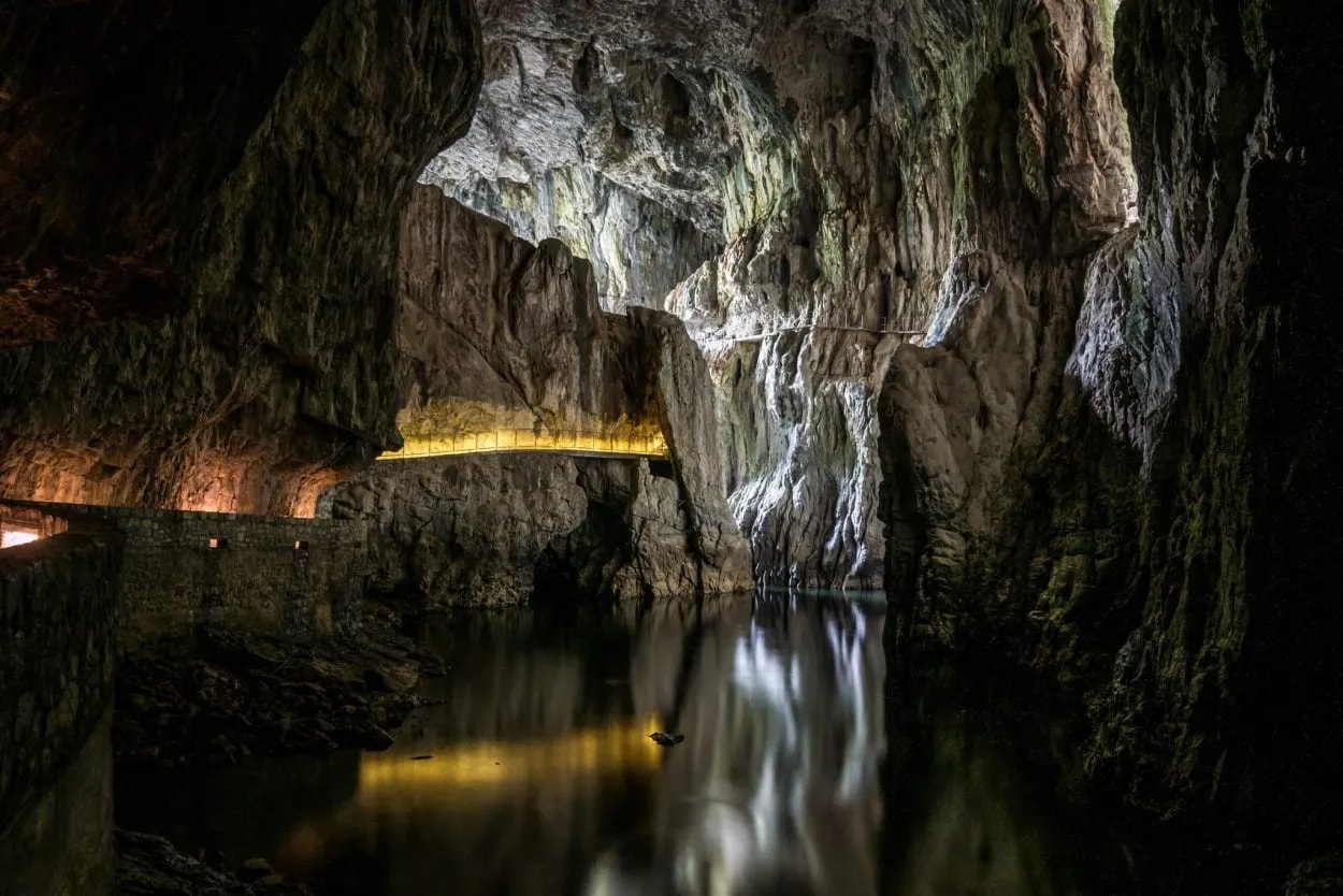 Underground river in Škocjan Caves, Slovenia, with illuminated walkway reflecting on dark water.