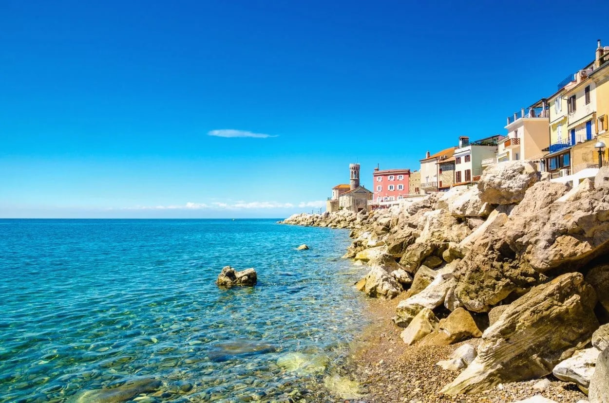 Rocky coastline and clear turquoise water next to colorful buildings in Piran, Slovenia.