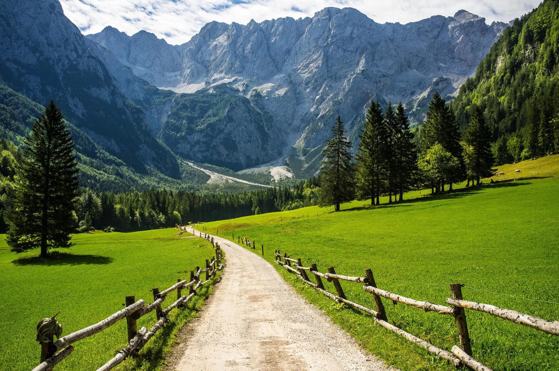 Dirt path with rustic fence leading toward massive gray mountains and green alpine meadows in Logar Valley.