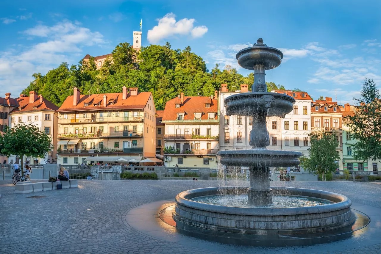Fountain in square with Ljubljana Castle on wooded hill and colorful buildings.