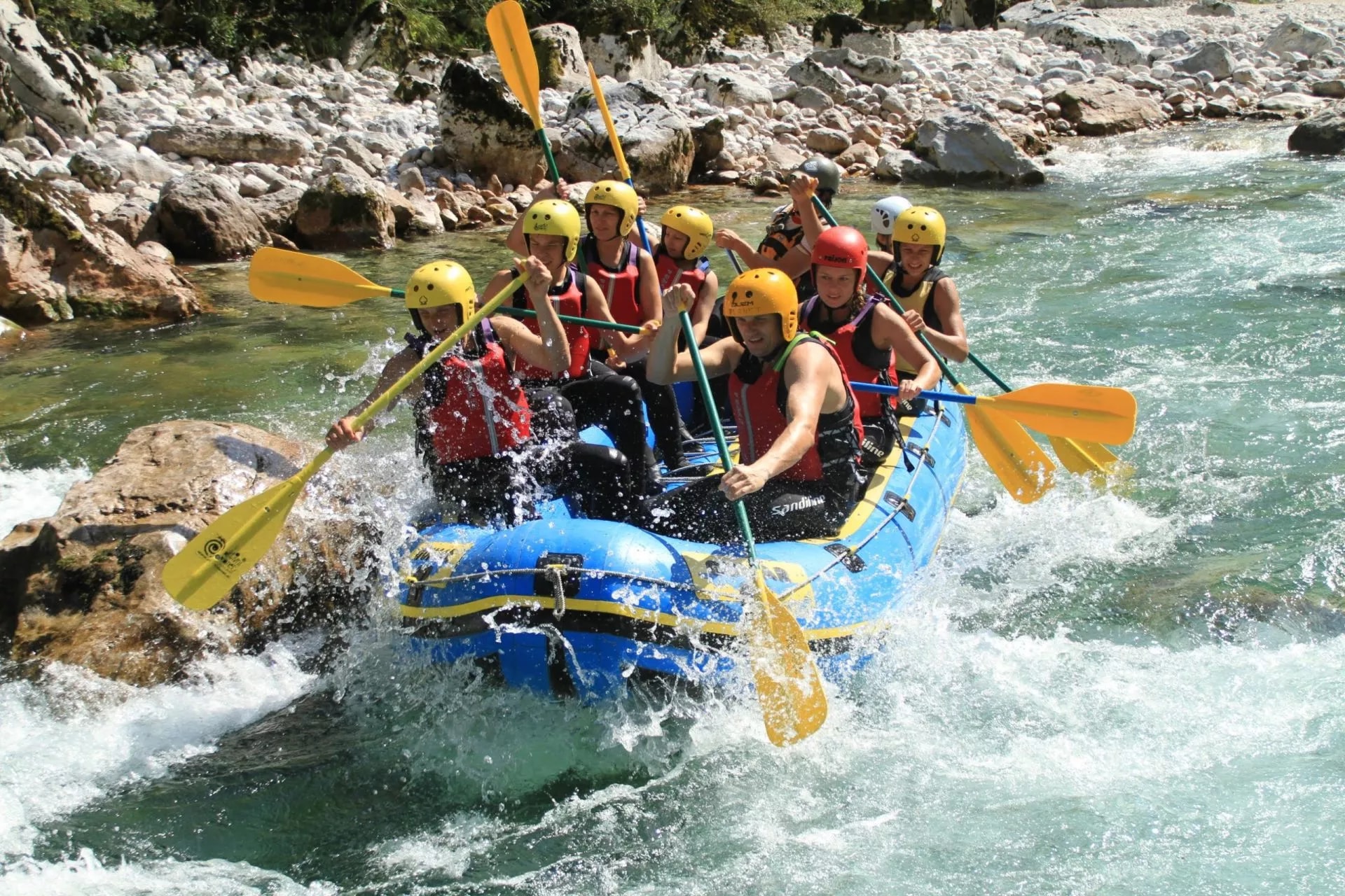 White water rafting group paddling blue raft through rapids near rocky river banks, Soca River.