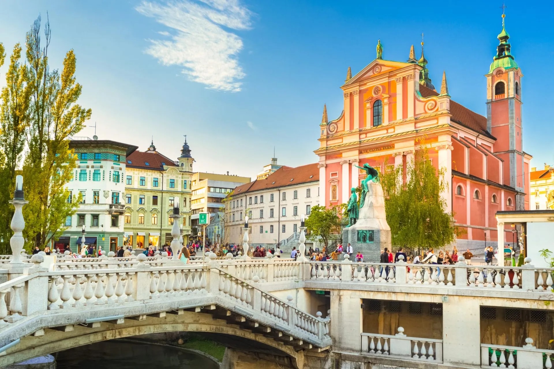 Triple Bridge (Tromostovje) and pink Franciscan Church in Ljubljana with people on the bridge.
