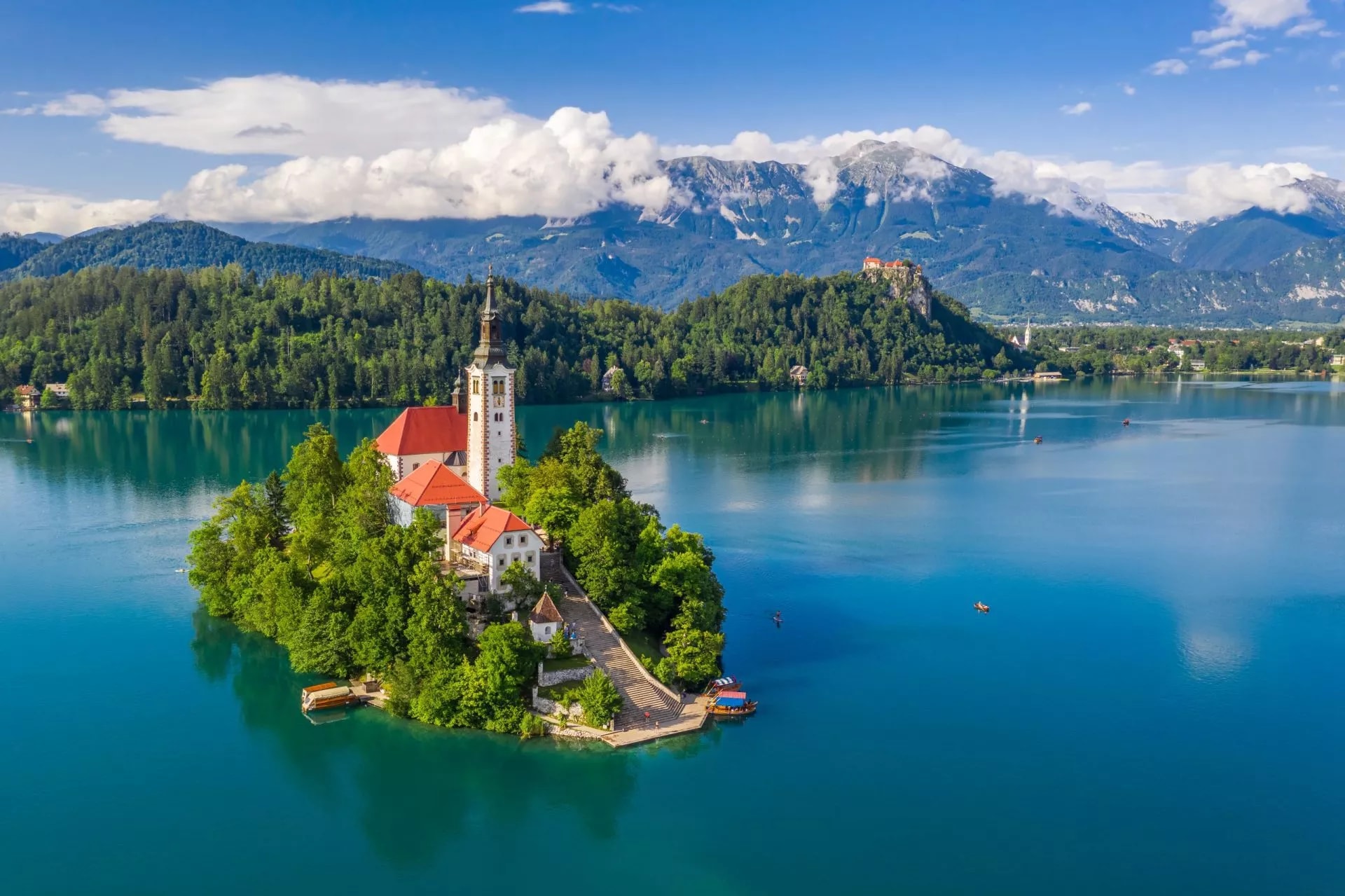 Lake Bled island church surrounded by turquoise water, mountains, and forest under blue sky.