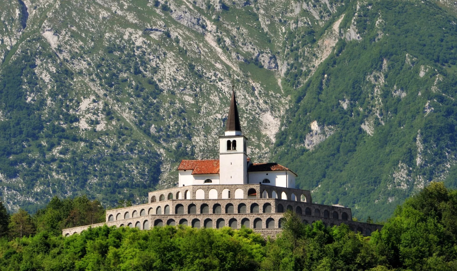White church with bell tower on arched stone base against lush green mountain slope.