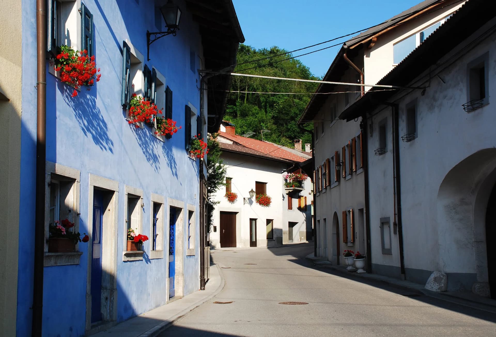 Buildings in Kobarid with bright blue facade and red geraniums line a sunny, narrow street.