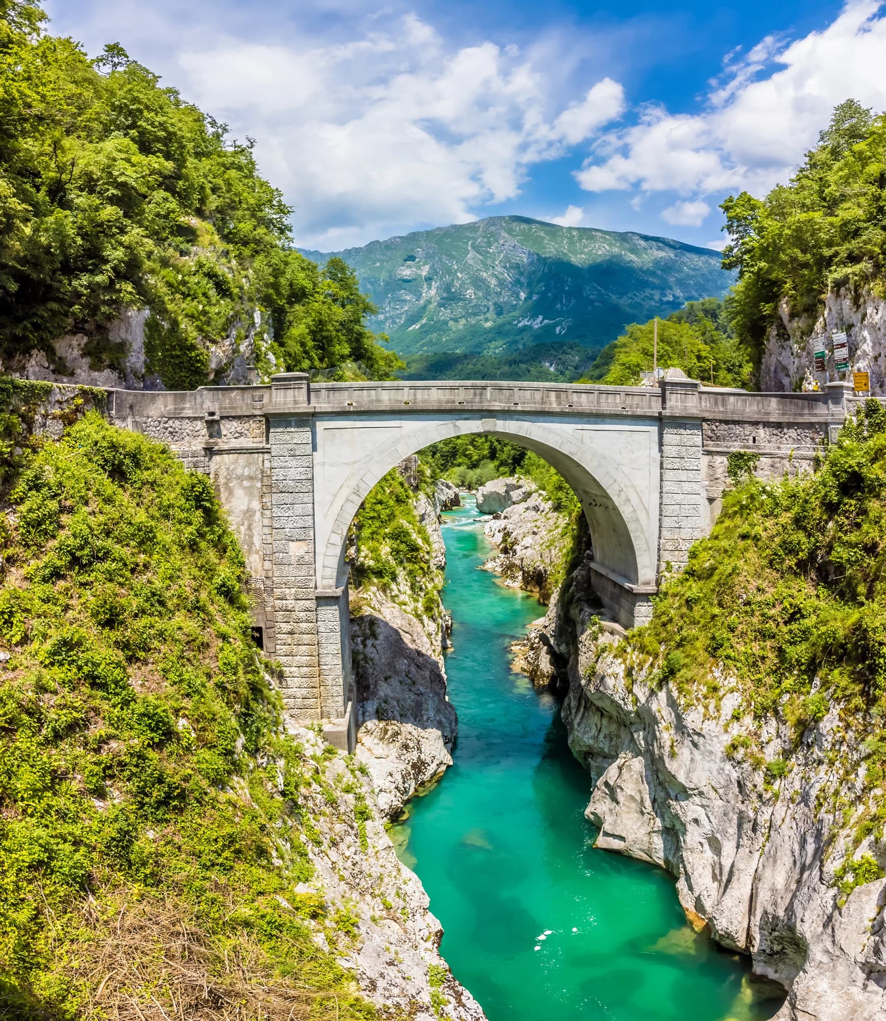 Napoleon Bridge over the turquoise Soca River gorge with lush green mountains under a blue sky.