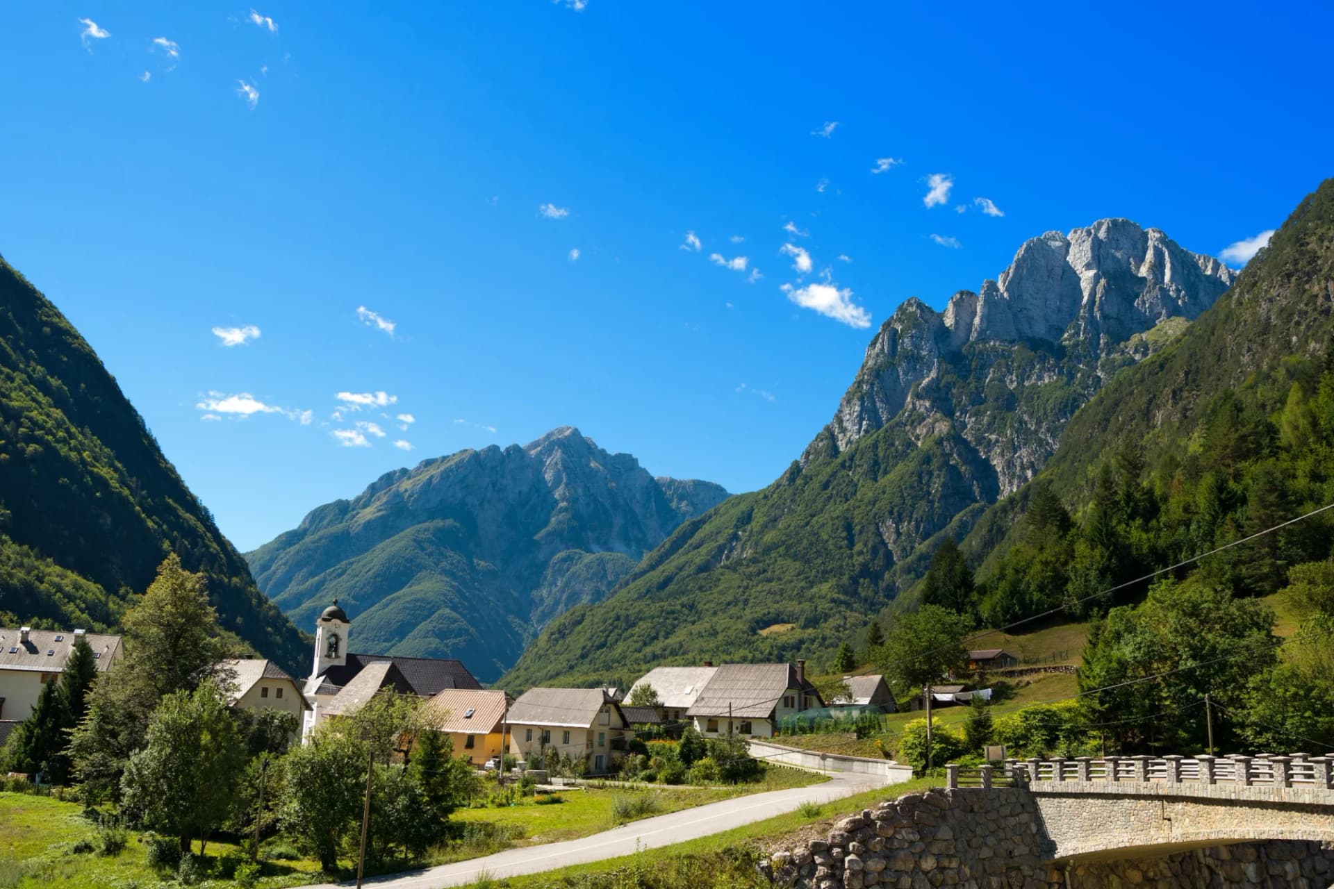Alpine village with church nestled between steep, forested mountains under a bright blue sky.