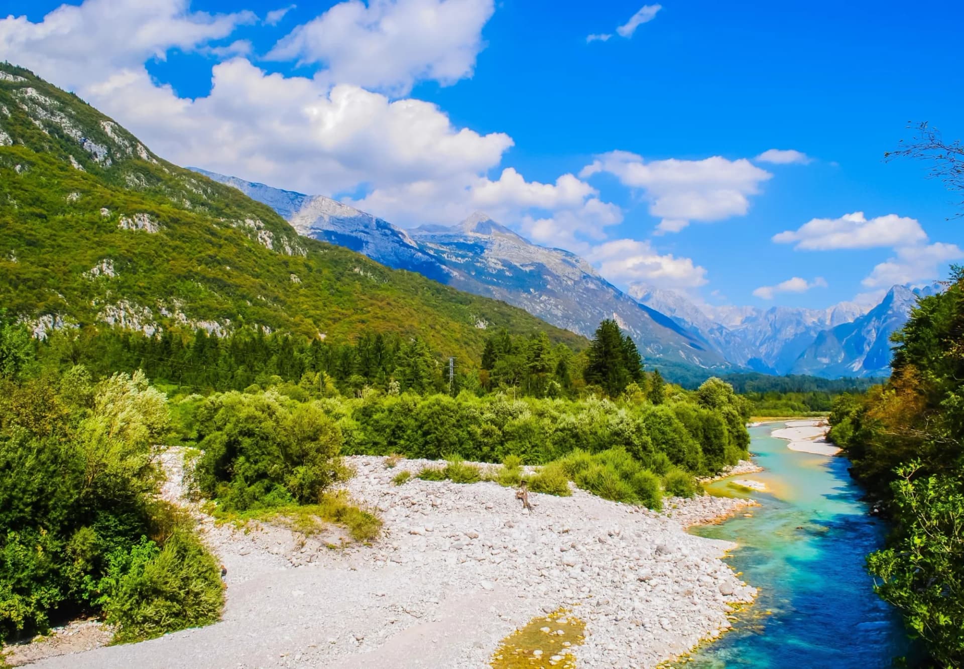 Mountain landscape with turquoise river, lush greenery, and rocky banks under a blue sky.