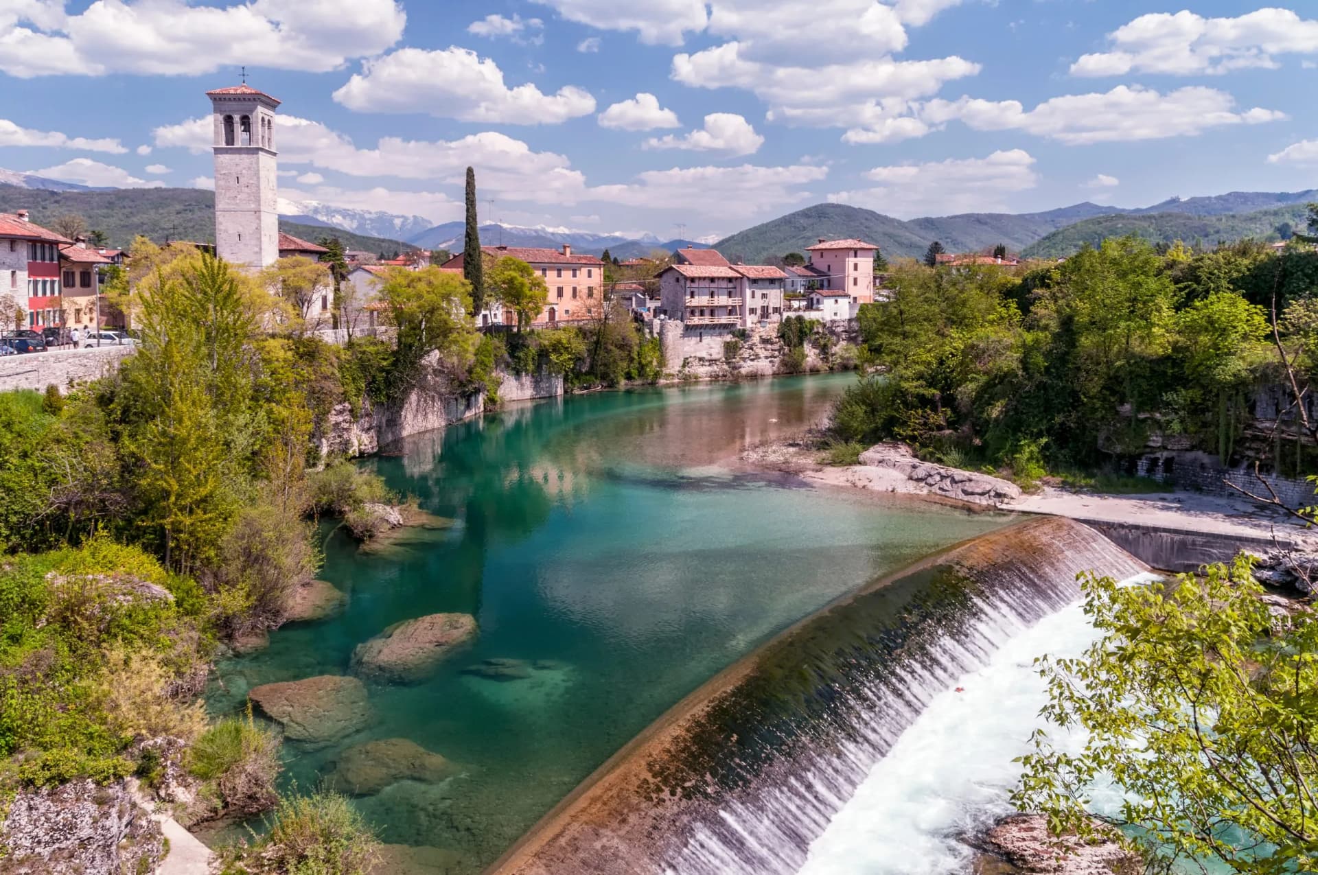 Cividale del Friuli town with bell tower, turquoise Natisone River waterfall, and snow-capped mountains.