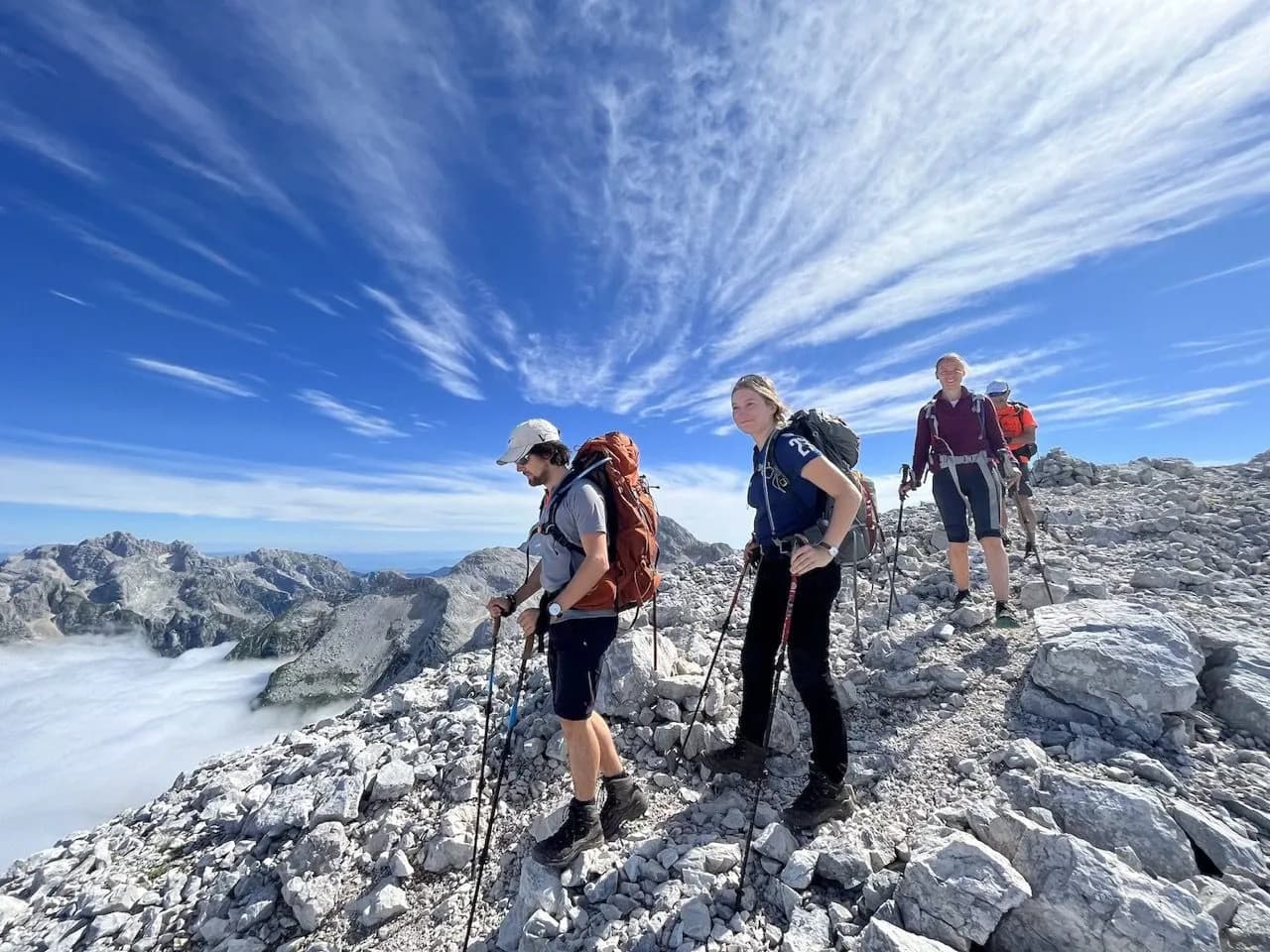Hikers with backpacks traverse a rocky summit above the clouds with Julian Alps visible.