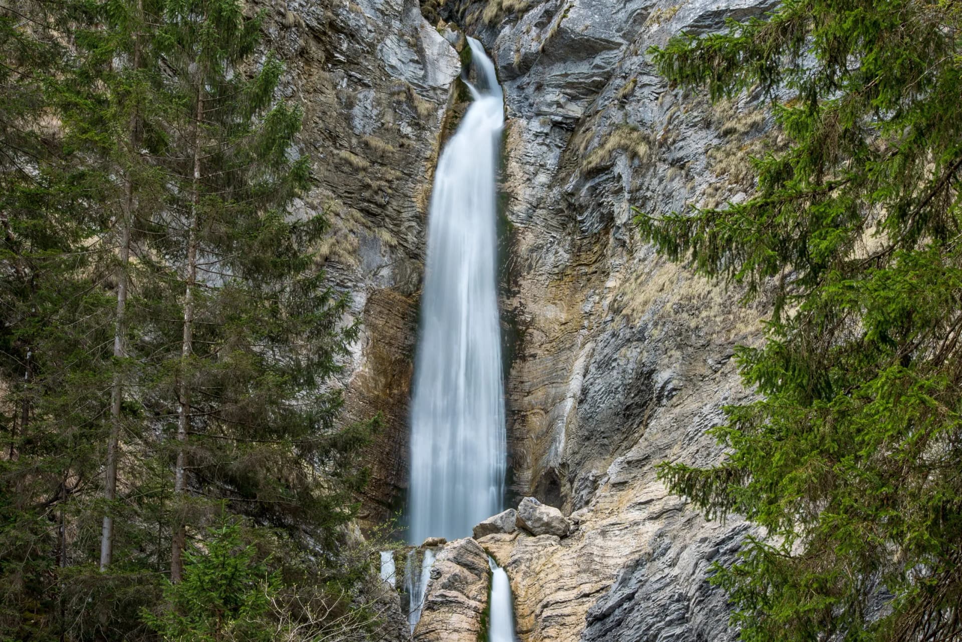 Tall waterfall cascading down rocky cliffs framed by green pine trees in Triglav National Park.