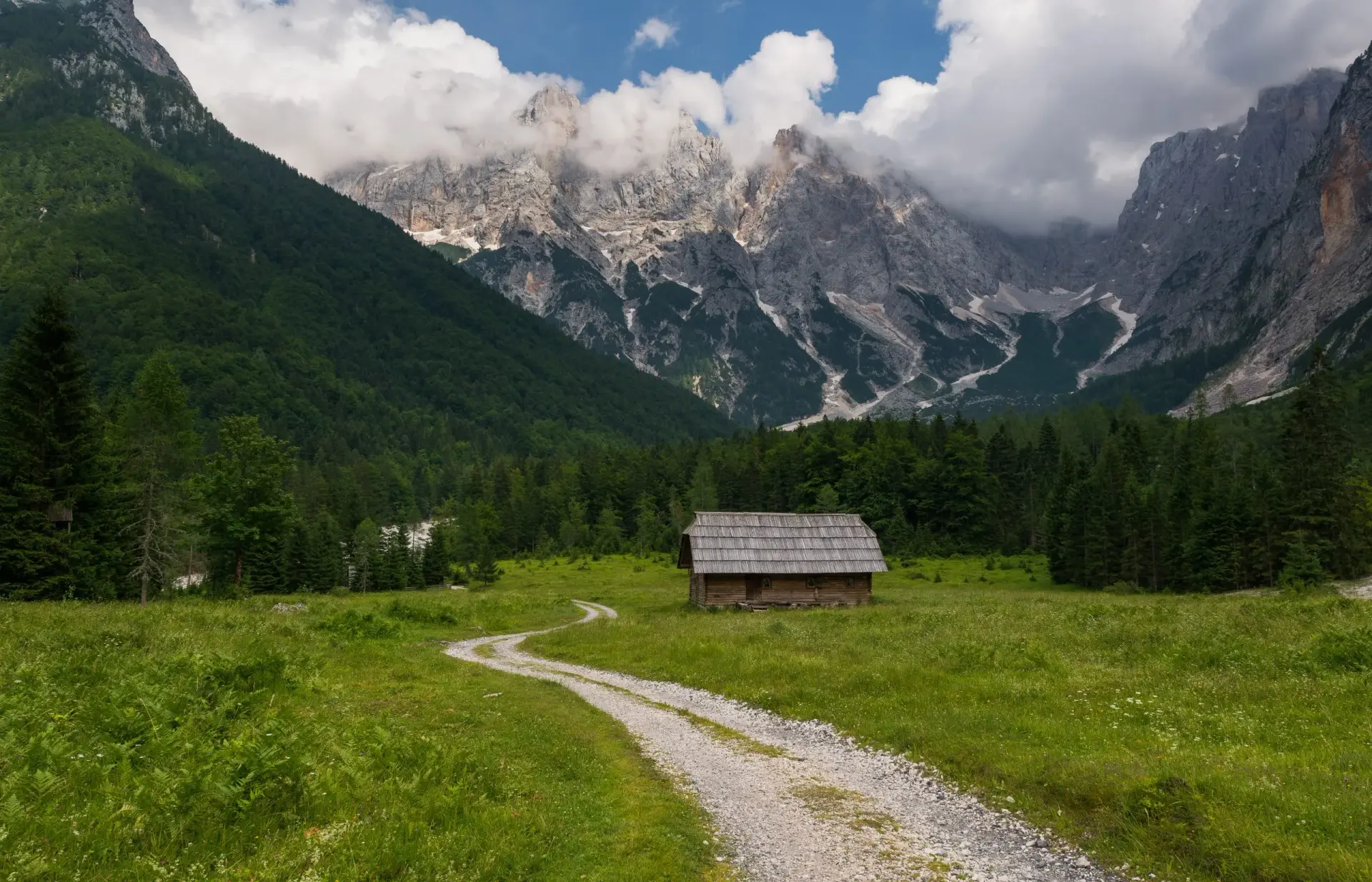 Wooden cottage in green meadow with gravel path, Julian Alps mountains in background