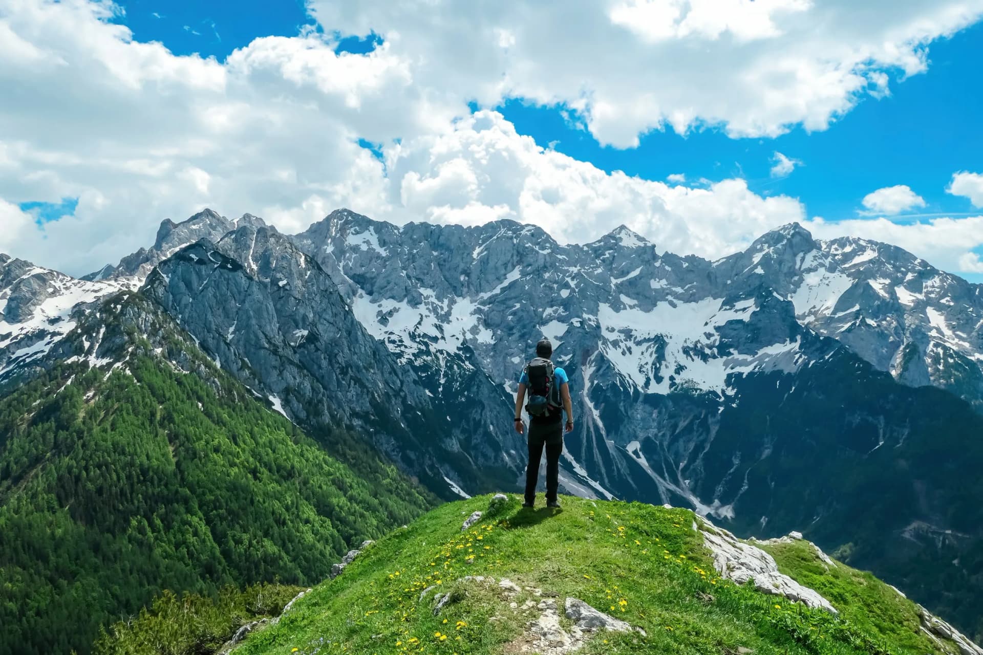 Hiker with backpack on grassy ridge overlooking snowy peaks of Kamnik-Savinja Alps.