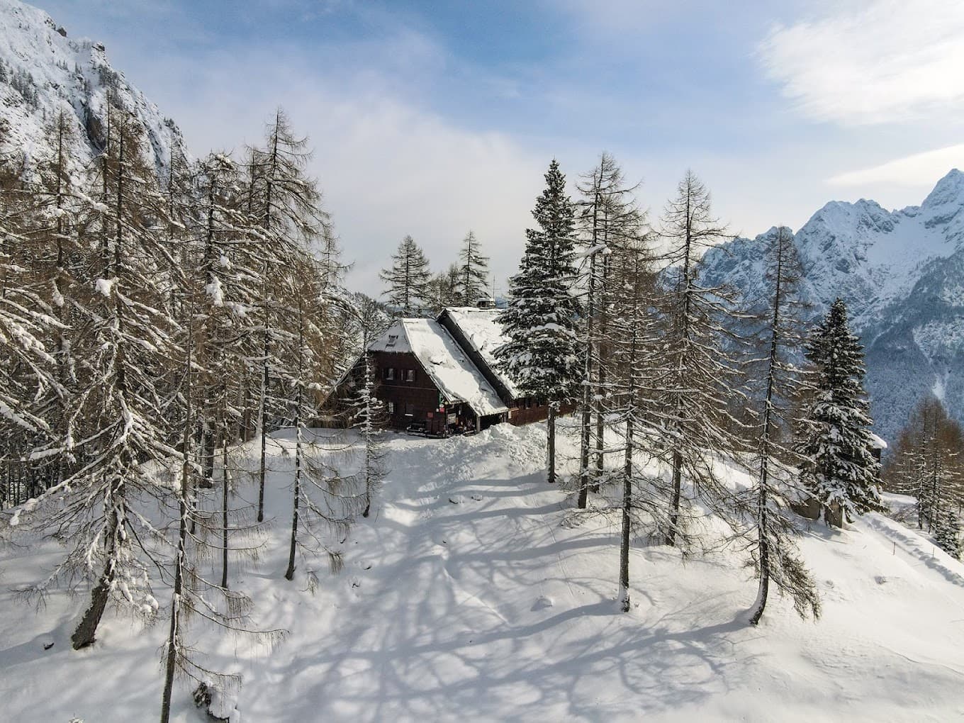 Mountain lodge covered in snow surrounded by snow-dusted pine trees and snowy peaks.