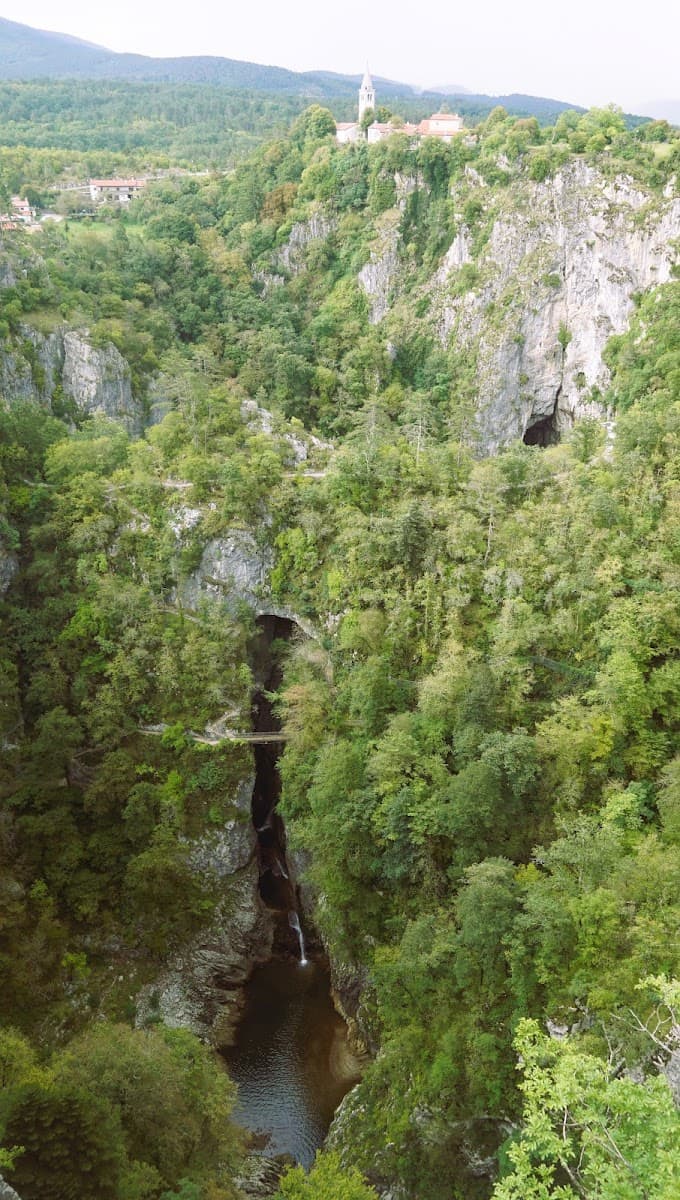 Deep gorge with waterfall, footbridge, and village with white church on clifftop.