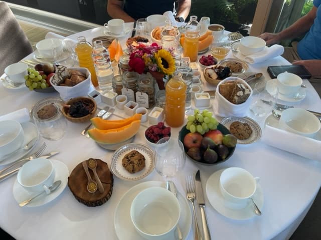 Abundant breakfast spread with fruit, bread, juice, and flowers on a white linen table.