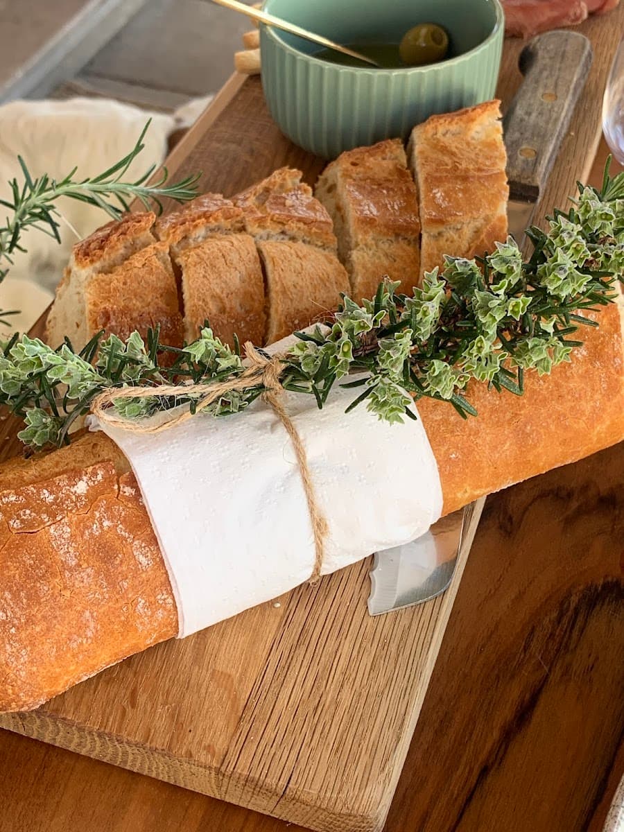 Sliced baguette wrapped in paper with herbs, served with olive oil on a wooden cutting board.