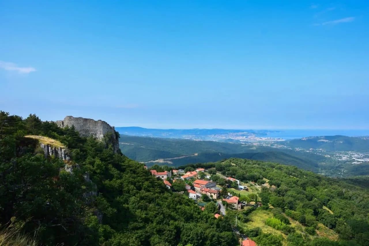 View from Socerb ruins over green hills, village, and distant coastline under a clear blue sky.
