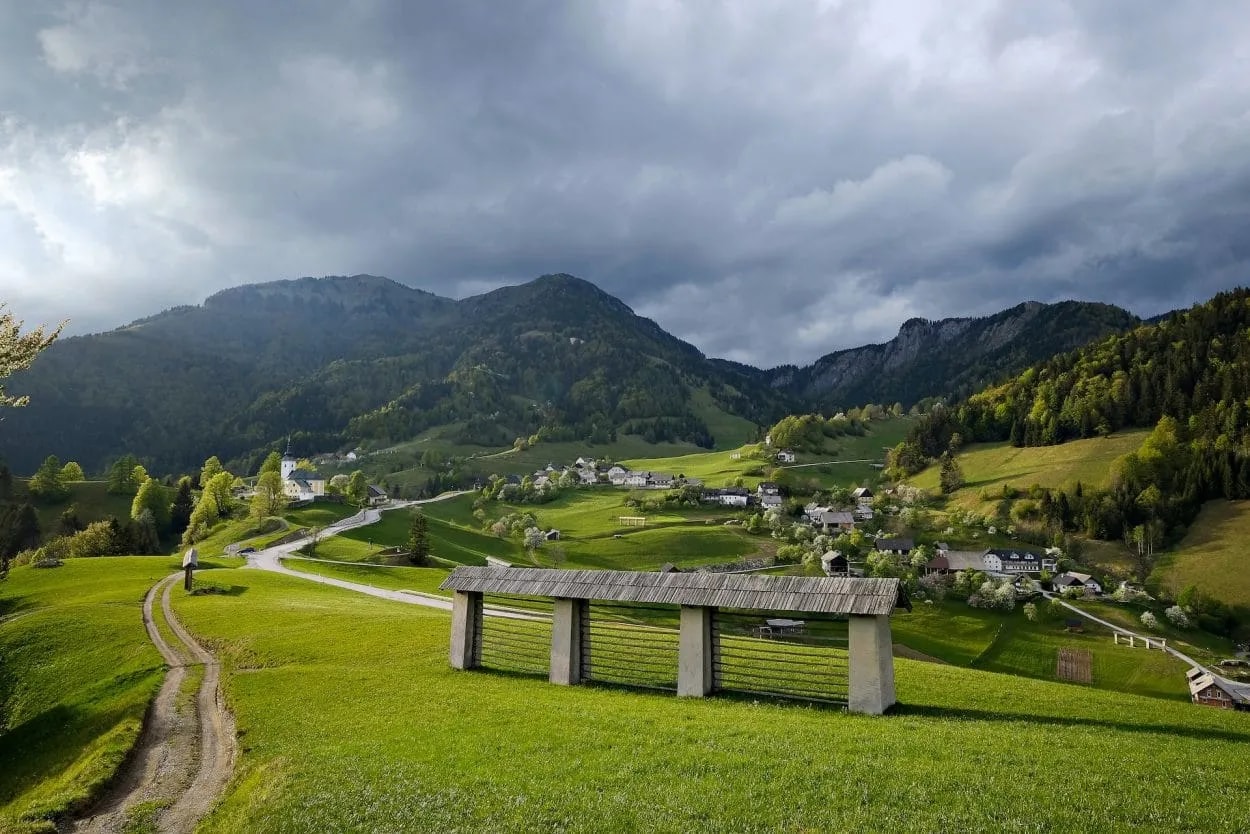 Scenic village of Sorica nestled in green hills with mountains under dark, dramatic clouds.
