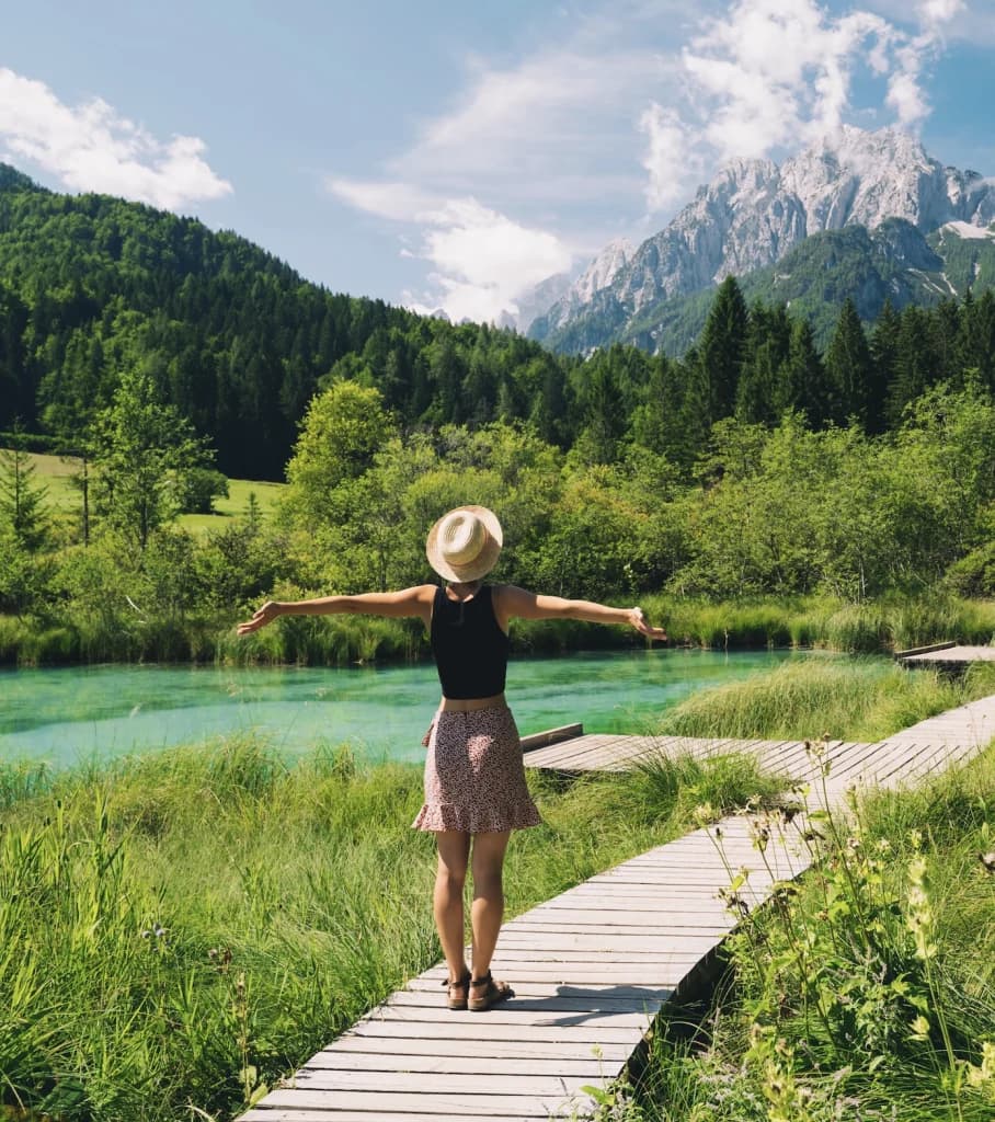 Woman in straw hat on boardwalk by turquoise water with forested mountains in background