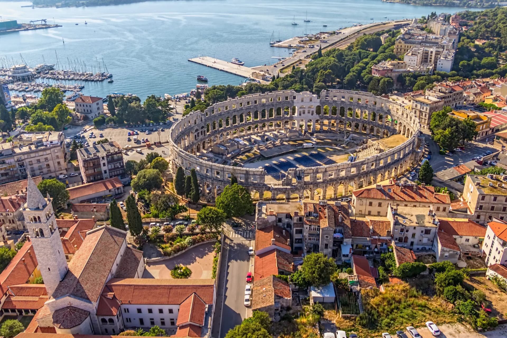 Aerial view of Pula Arena amphitheater overlooking harbor with docked sailboats and city buildings.