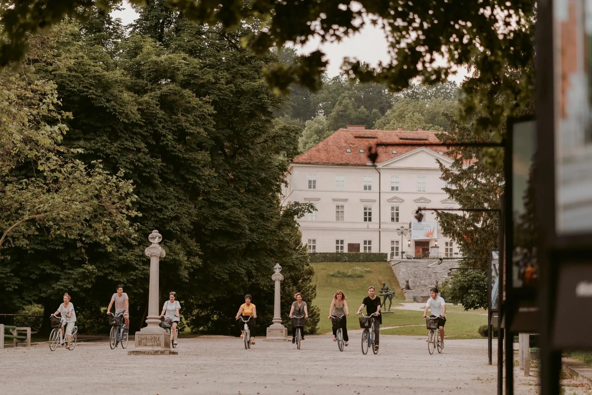 Cyclists riding bikes in Ljubljana's Tivoli Park past a historic building.