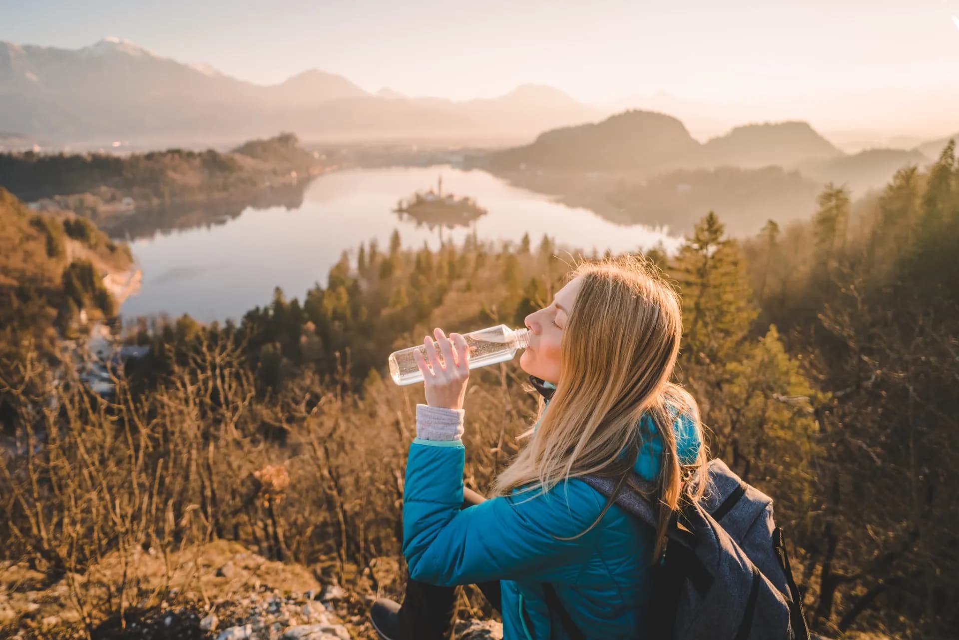 Hiker drinking water overlooking Lake Bled island and mountains at sunset