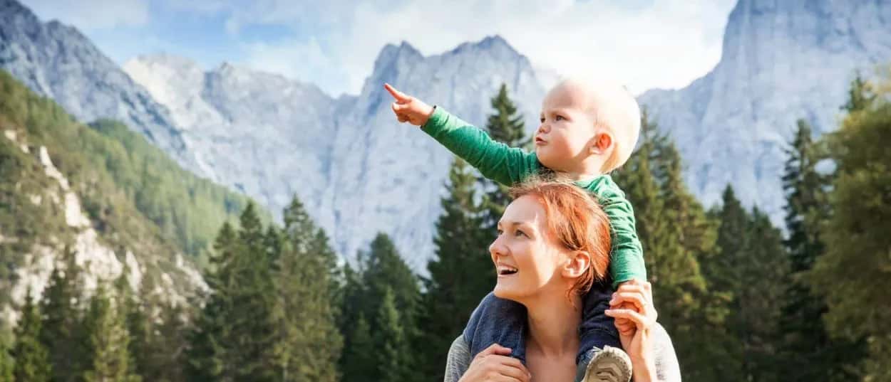 Mother carrying child on shoulders hiking near tall, rocky mountains and pine forest in Kranjska Gora.