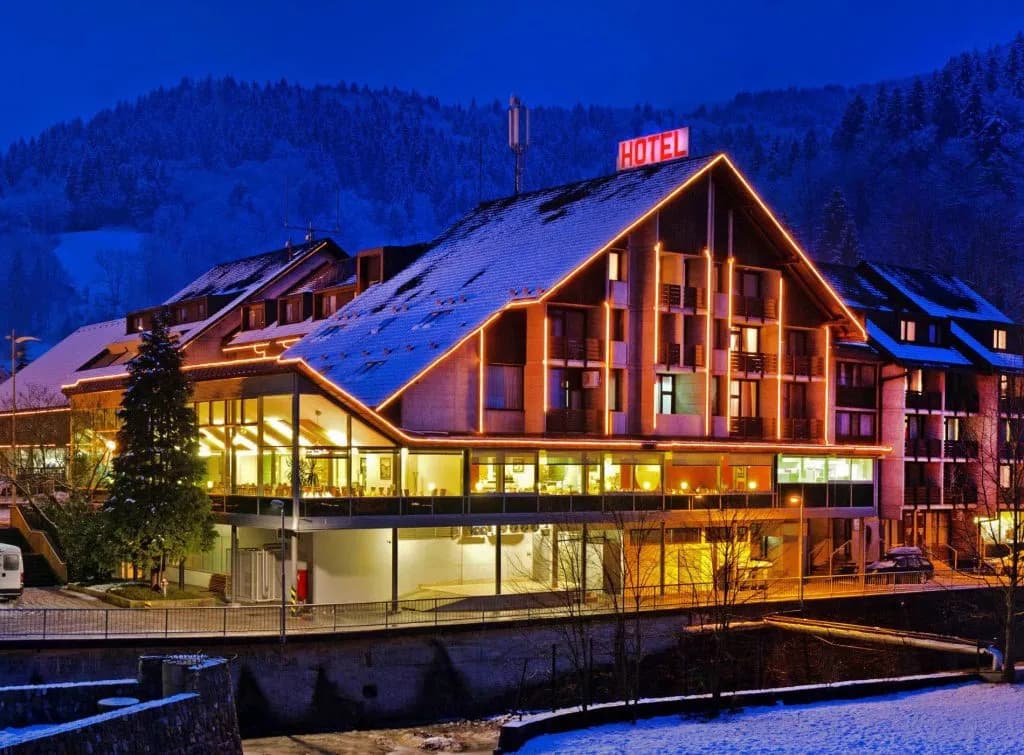 Hotel Cerkno in winter illuminated at night with snow-covered roof and forested mountains.