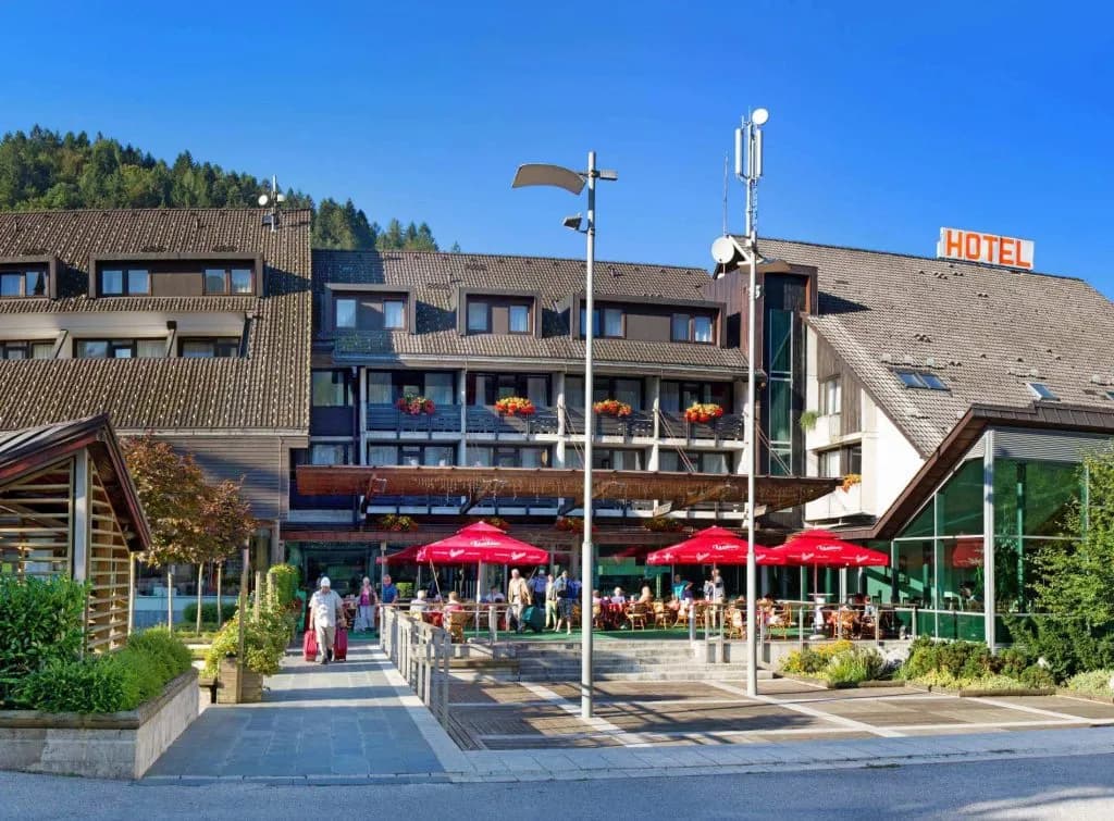 Hotel Cerkno terrace with red umbrellas and guests, set against a forested mountain backdrop.