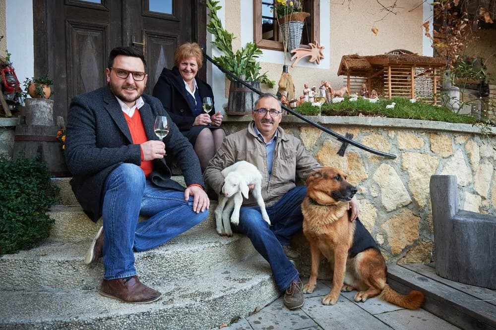Family posing on stone steps with wine, holding a lamb, next to a German Shepherd near a nativity scene.