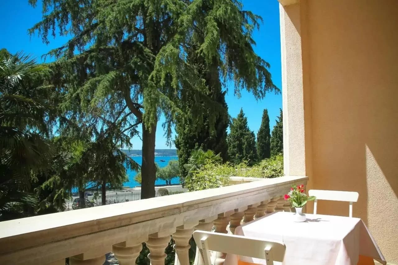 Balcony with table and chairs overlooking turquoise sea, lush trees, and clear blue sky.
