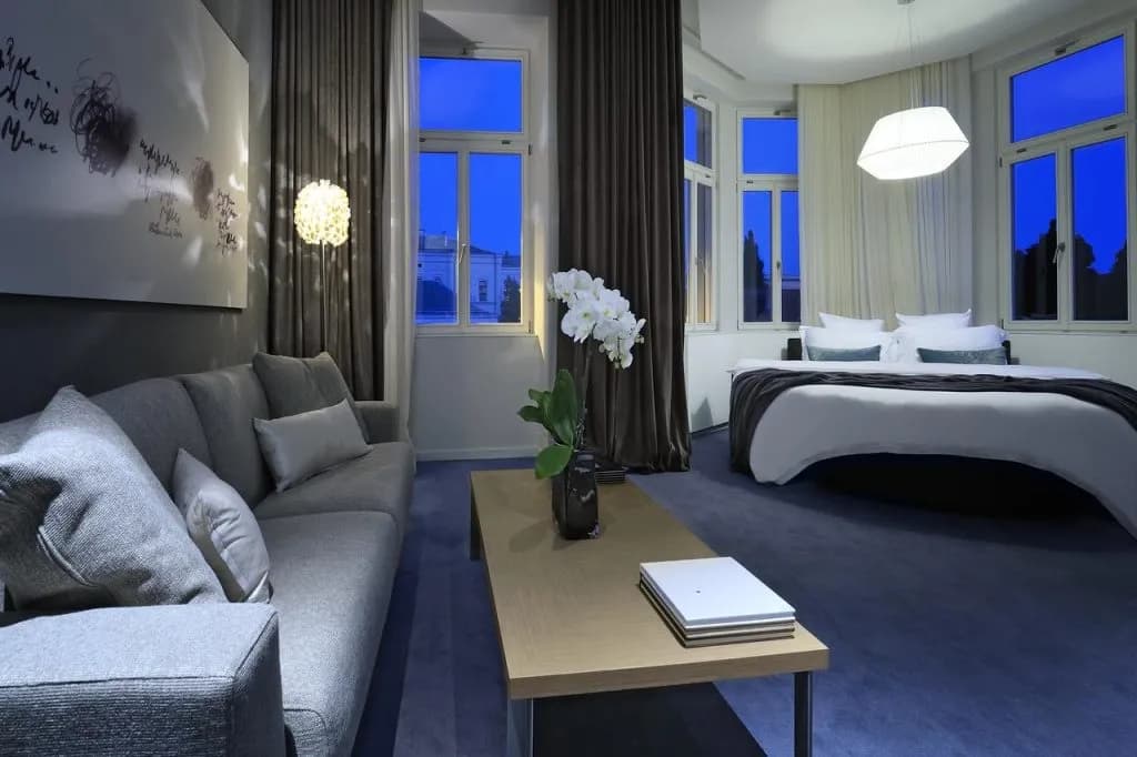 Modern hotel room with gray sofa, wood coffee table, white orchid, and large bed at twilight.