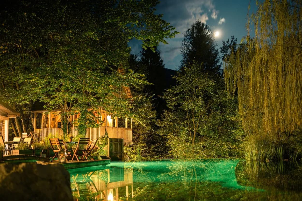 Wooden dining area by glowing green water at night in Garden Village Bled, Slovenia.