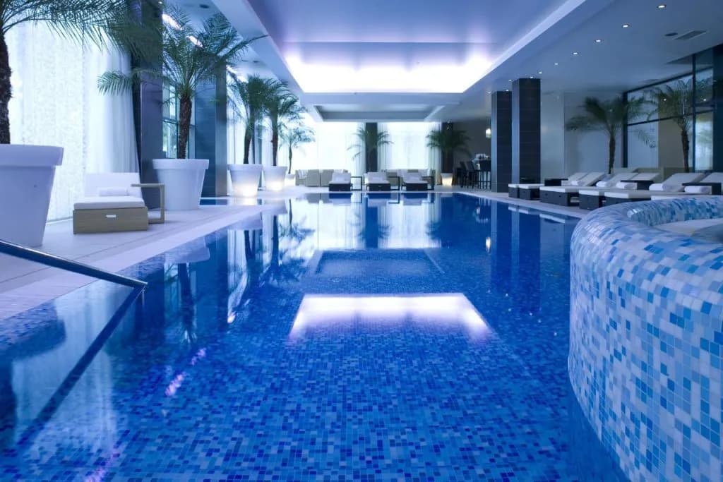 Indoor swimming pool with blue mosaic tiles, illuminated ceiling, and potted palm trees at Hotel Kempinski Palace.